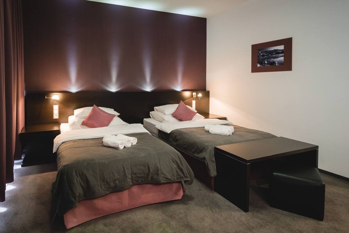 Quarto de hotel com duas camas de solteiro, uma mesa de cabeceira, uma escrivaninha e uma cadeira.