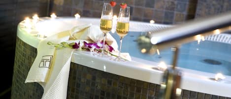 A hot tub with candles, two glasses of champagne, and a towel on a tiled surface.