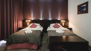 Una habitación de hotel con dos camas, cada una con cabecero, almohadas y toallas. Hay mesitas de noche y una lámpara fijada a la pared.