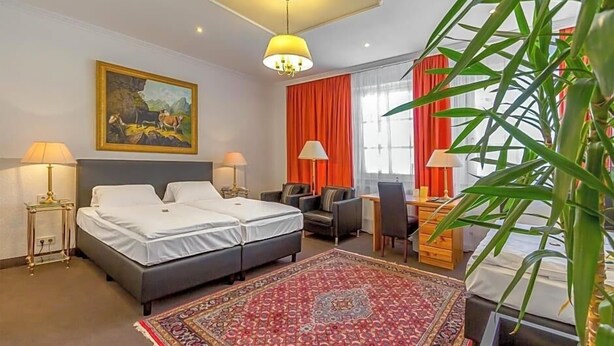 A hotel room with two beds, a desk, a chair, a red rug, a painting, and a large window with curtains. - Hotel Adler (Ingolstadt)