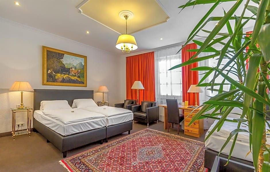 Une chambre d’hôtel avec deux lits, un bureau, une chaise, un tapis rouge, un tableau et une grande fenêtre avec des rideaux.