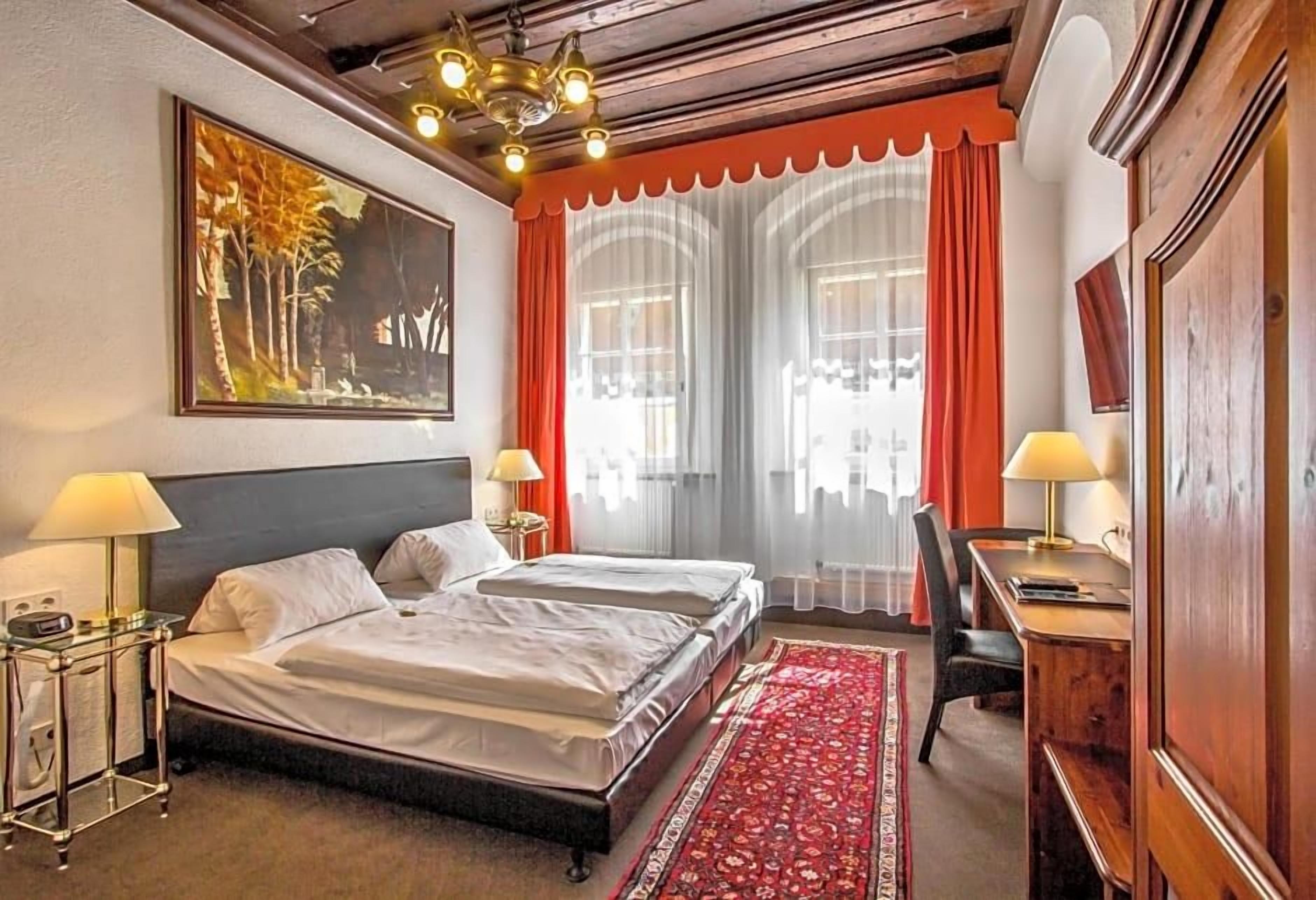 A hotel room with a double bed, two bedside tables with lamps, a desk with a chair, a large window with curtains, and a painting on the wall.