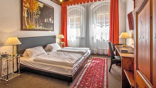 A hotel room with a double bed, two bedside tables with lamps, a desk with a chair, a large window with curtains, and a painting on the wall. - Hotel Adler (Ingolstadt)
