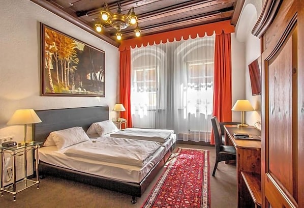 A hotel room with a double bed, two bedside tables with lamps, a desk with a chair, a large window with curtains, and a painting on the wall. - Hotel Adler (Ingolstadt)
