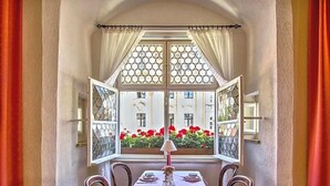 A dining area with a table set for four, a window with a view of buildings and red flowers, and curtains on either side. - Hotel Adler (Ingolstadt)