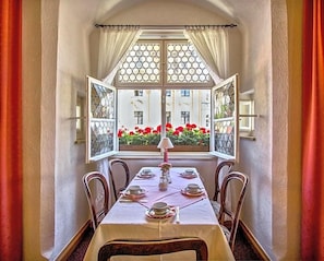 A dining area with a table set for four, a window with a view of buildings and red flowers, and curtains on either side. - Hotel Adler (Ingolstadt)