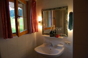 A bathroom with a white sink, a mirror, and a window with curtains. - Hotel Schweizerhof (Wildschönau)