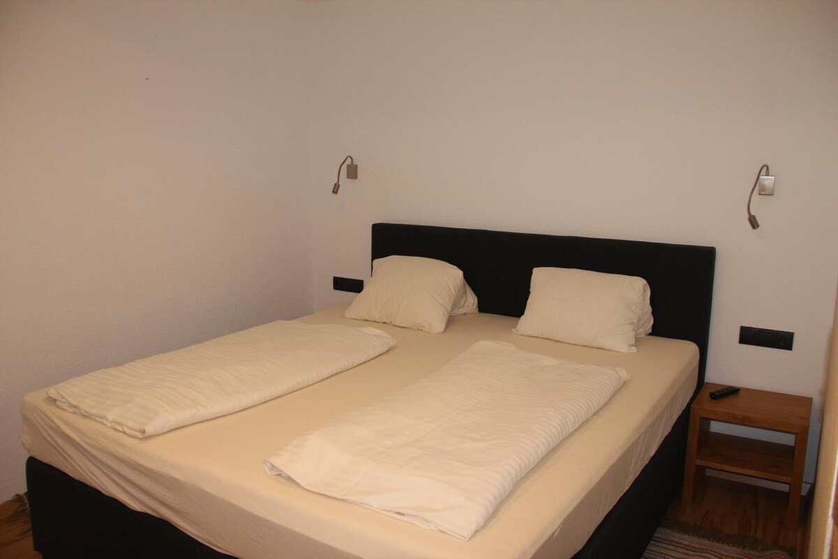 Un letto singolo con una testiera nera, due cuscini e una coperta. Un comodino con un telecomando sopra.