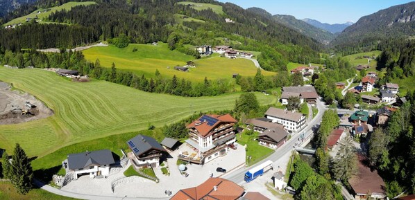 A mountainous village with a central building surrounded by residential houses, a main road, and green fields.