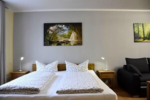 Una cama doble con cubrecama floral, dos mesitas de noche con lámparas, un sofá negro y dos cuadros colgados en la pared con paisajes naturales.