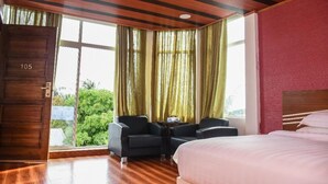 A hotel room with a large bed, a small table with two chairs, a wooden door with a number, and a view of greenery through the window. - Sunset Stay Maldives (Thinadhoo)