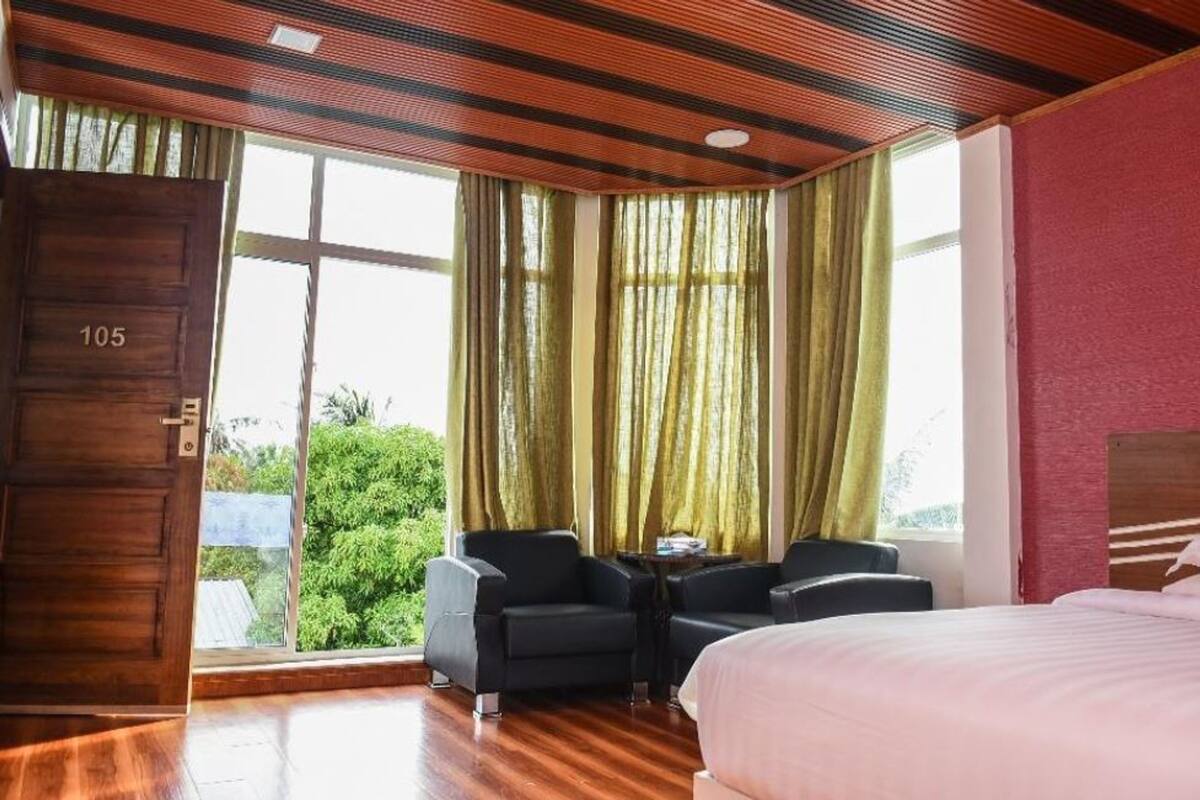 A hotel room with a large bed, a small table with two chairs, a wooden door with a number, and a view of greenery through the window.