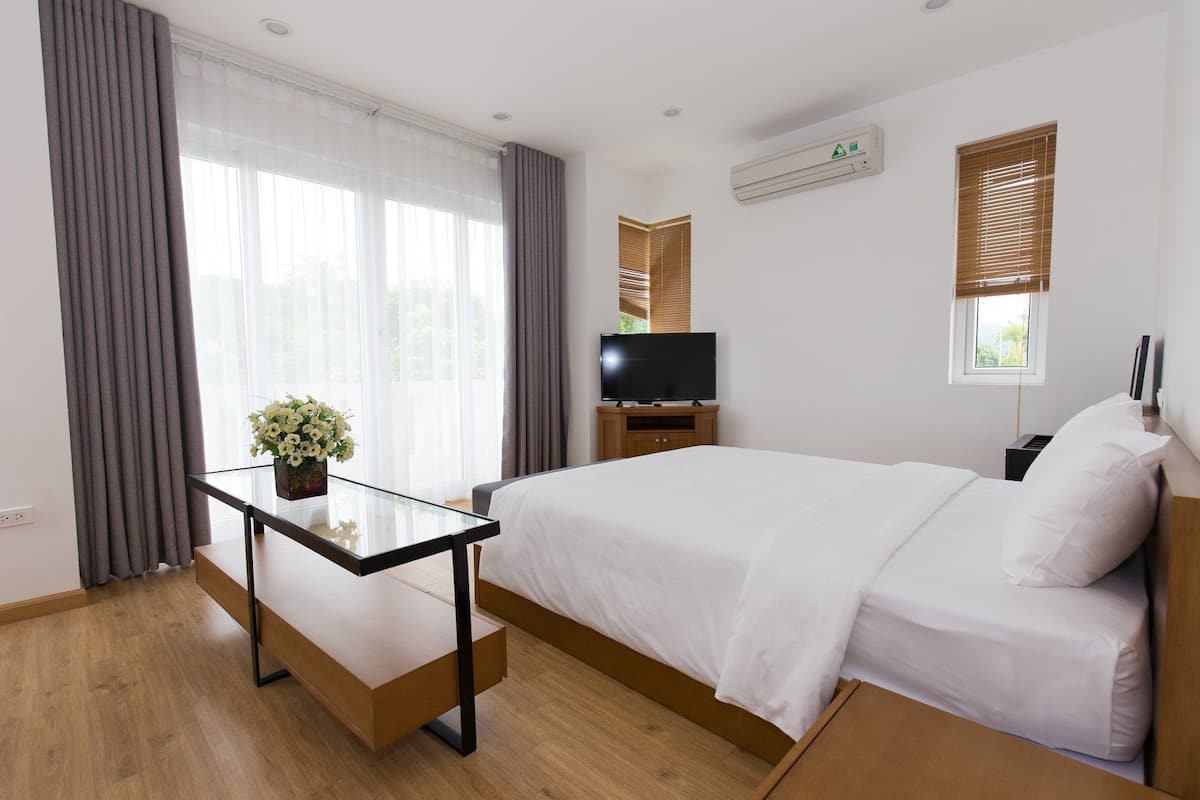 Ein Hotelzimmer mit Bett, Fernseher, Schreibtisch mit Pflanze und Fenster mit Jalousien.