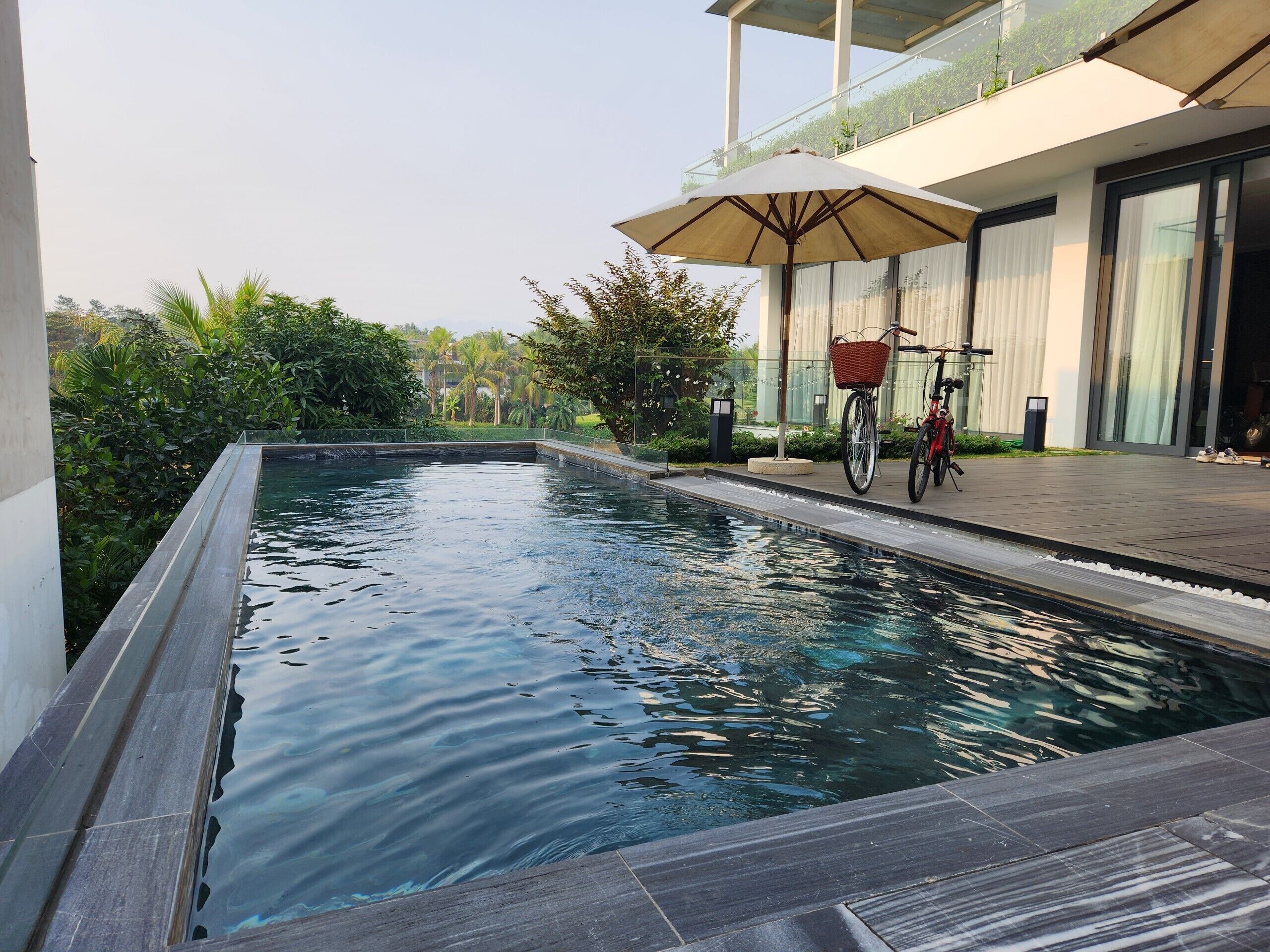 A swimming pool with a bicycle and a basket, surrounded by greenery and a modern building.