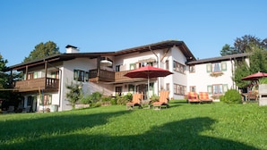 A two-story building with a balcony, outdoor seating with red umbrellas, and a well-maintained lawn. - Ferienwohnungen Zum Sommerfrischler (Seehausen am Staffelsee)