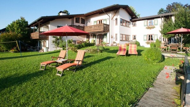 A two-story building with a balcony, a grassy yard, and outdoor seating with umbrellas. - Ferienwohnungen Zum Sommerfrischler (Seehausen am Staffelsee)