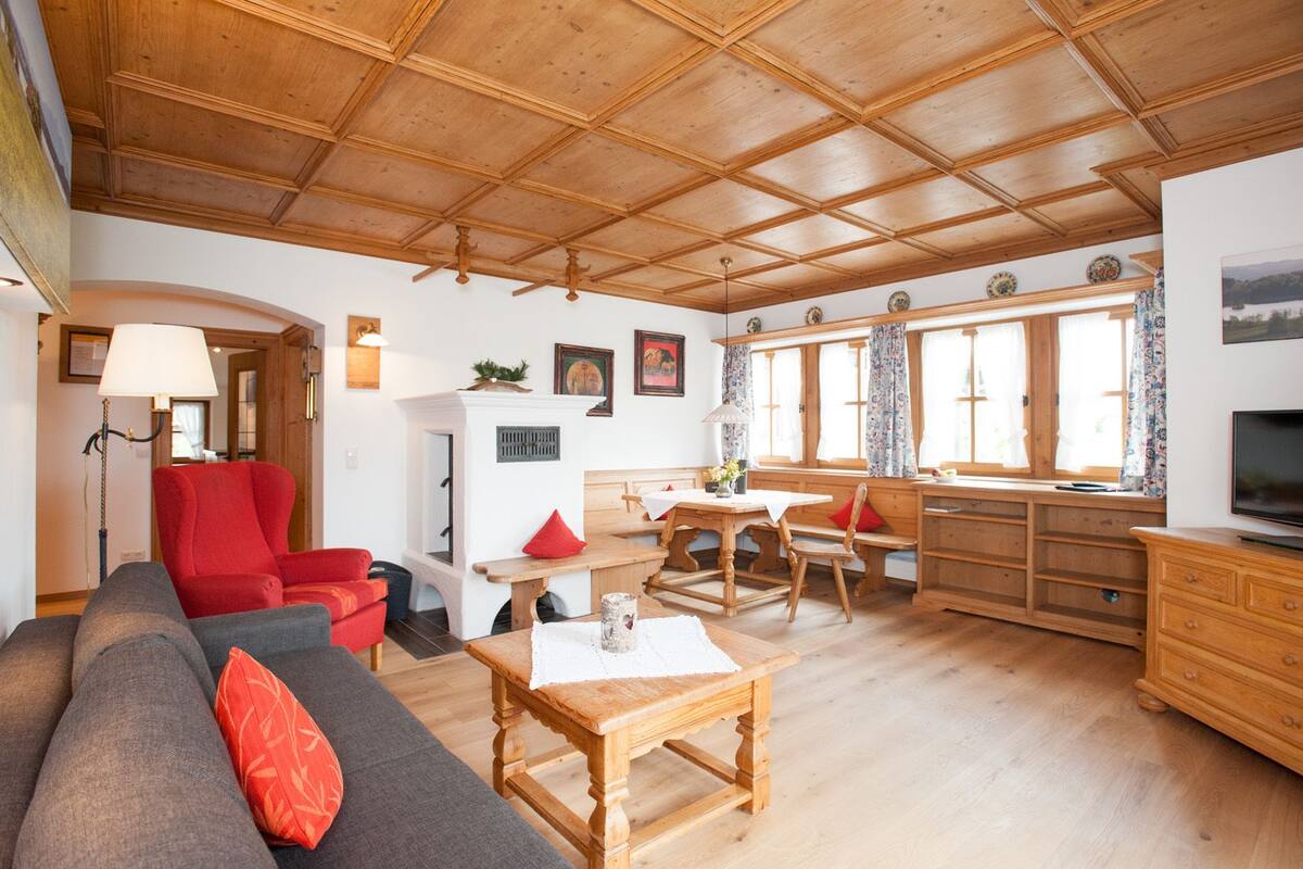 A cozy living room with a wooden ceiling, a sofa, a red armchair, a dining table, and a TV.