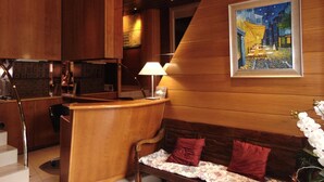 A hotel lobby with a wooden reception desk, a sofa with red cushions, a painting of a café scene, and a staircase. - Hostal Piteus (Sant Llorenc de Morunys)