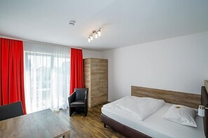 A hotel room with a bed, a chair, a table, and a window with red curtains. - Come In Hotel (Ingolstadt)