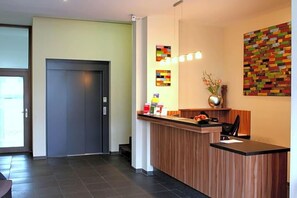 A reception desk with a wooden counter, a black chair, and a vase with flowers. There are framed pictures on the wall and a door to the left.