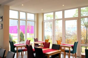 A room with large windows, multiple tables, and chairs arranged for dining.