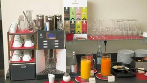 Una barra de desayuno con máquina de café, dispensadores de té y café, una variedad de tazas y una selección de alimentos para el desayuno.
