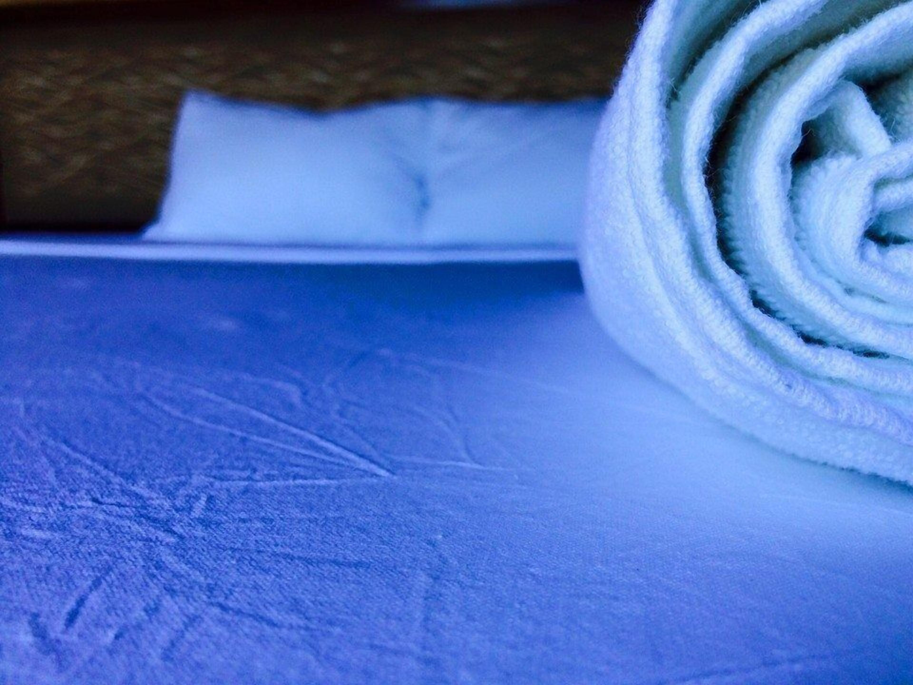 Une serviette blanche enroulée, posée sur une surface bleue texturée, qui pourrait être un lit ou une table.