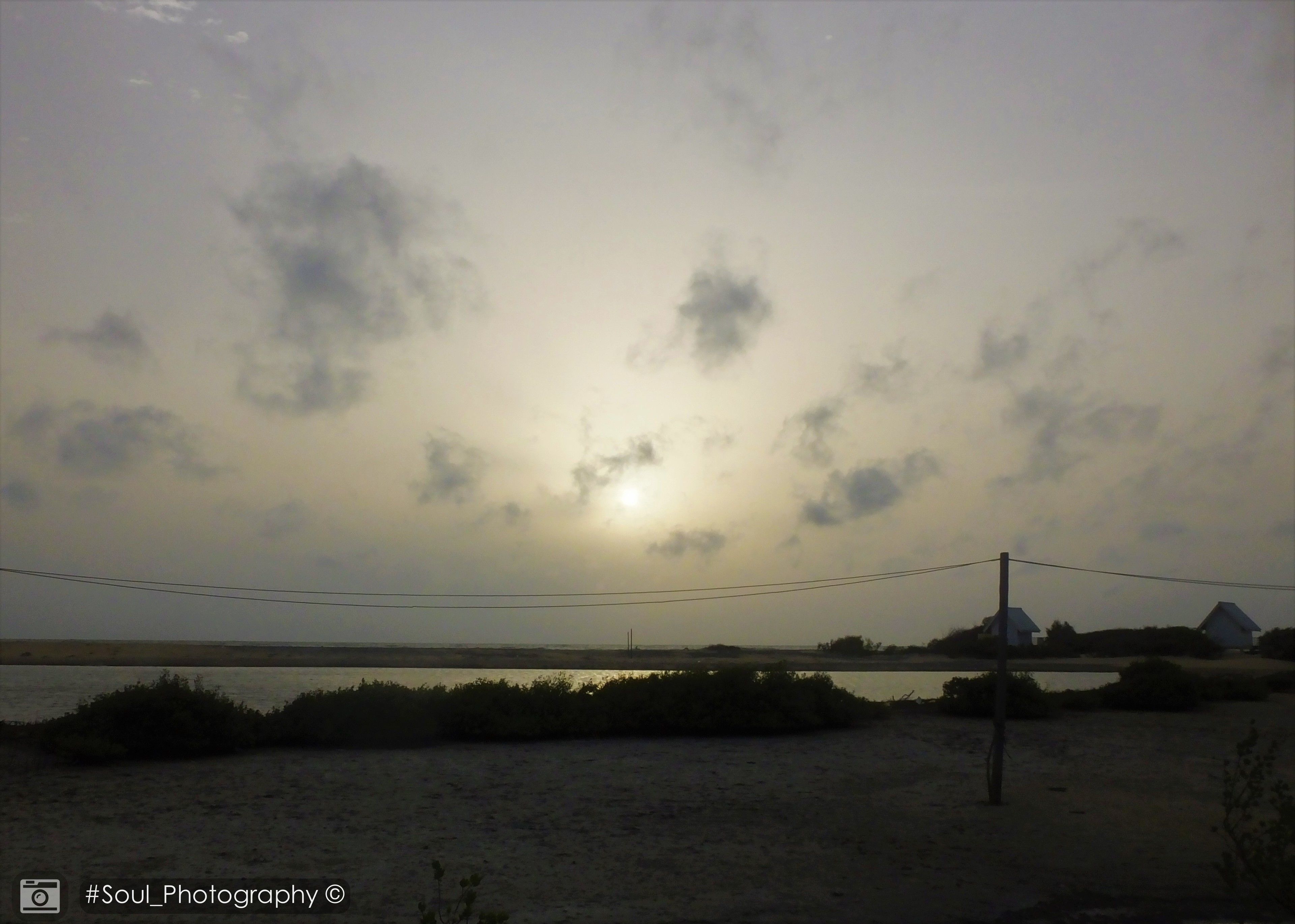 A beach with a cloudy sky and a body of water. There are power lines and a few buildings in the background.