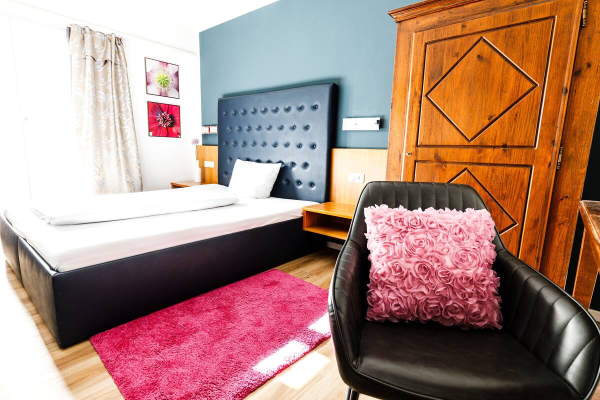Een slaapkamer met een bed, een houten kledingkast, een zwarte leren fauteuil met een roze kussen met bloemenmotief, en een rood vloerkleed.