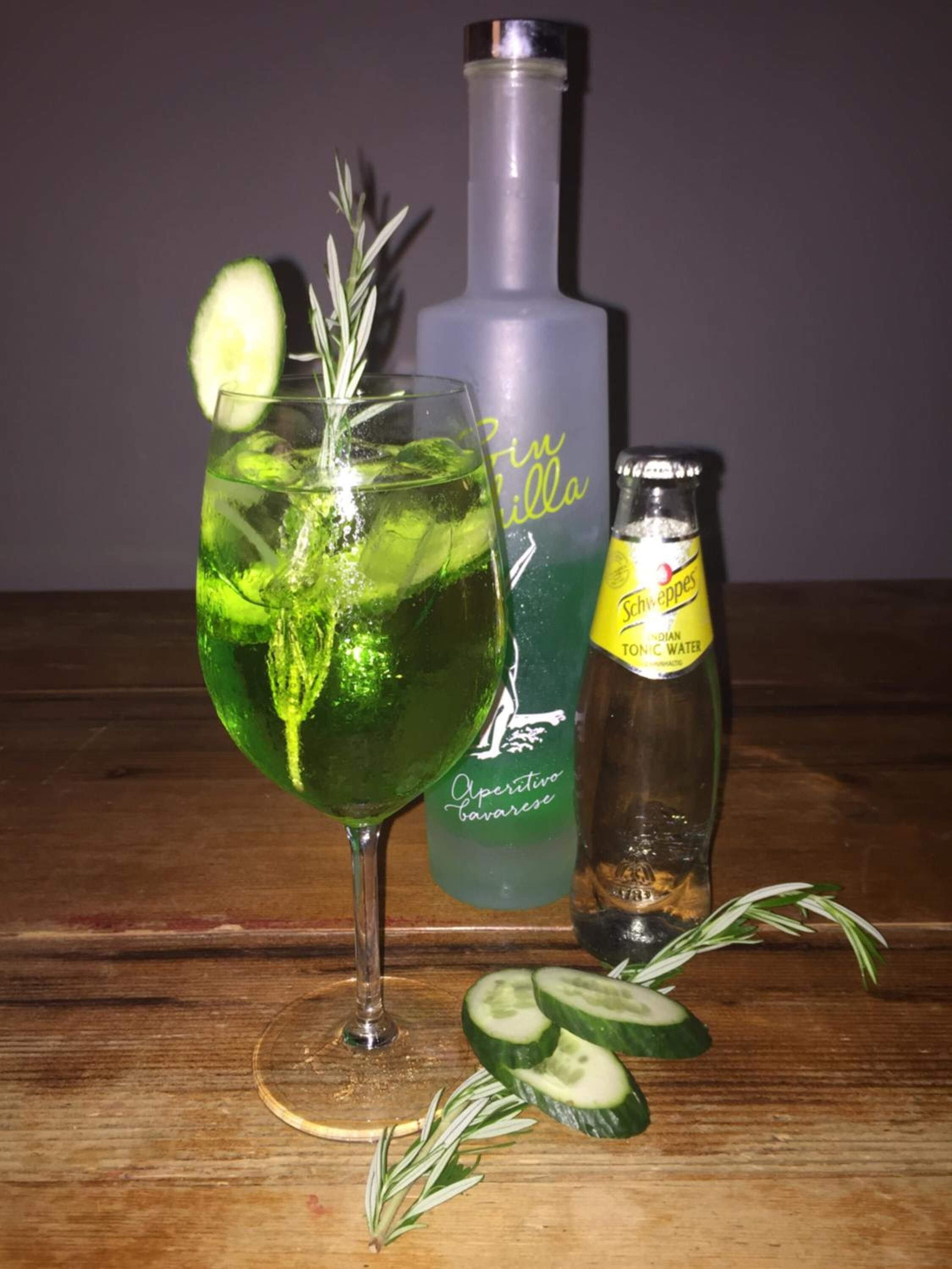 A glass of green drink with cucumber slices and rosemary, a bottle of Schweppes tonic water, and a bottle of Aperitivo Bavarese on a wooden table.