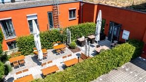 An outdoor patio with tables and chairs, surrounded by greenery and a building with orange walls.