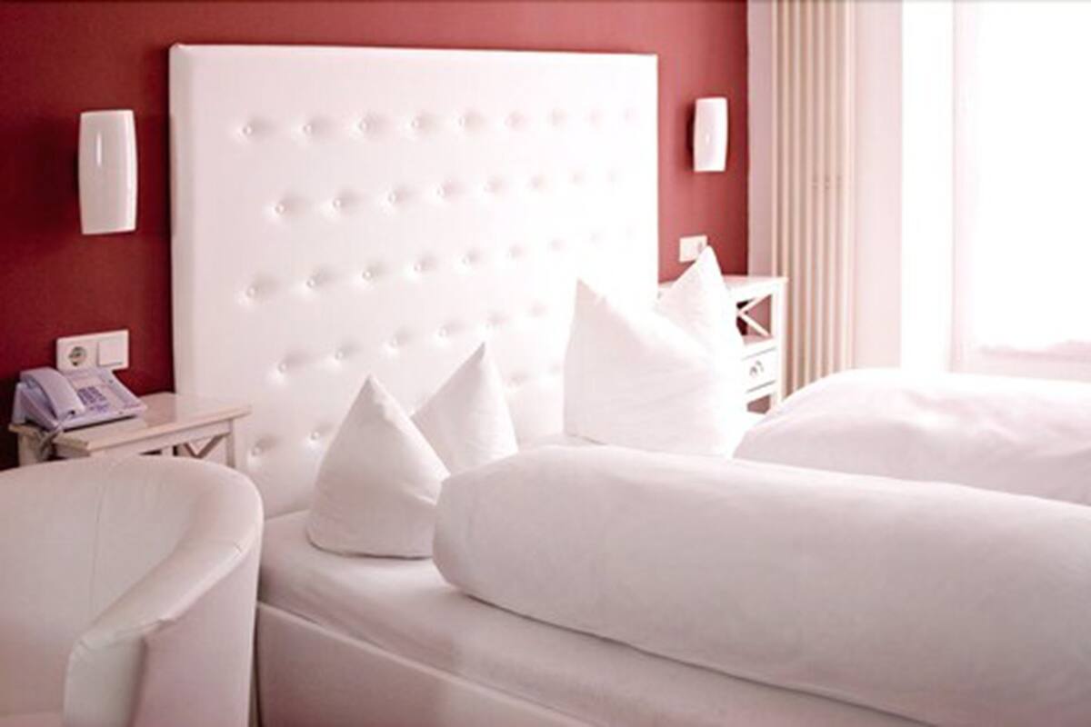 Een hotelkamer met een groot wit hoofdbord, twee bedden, witte kussens, een nachtkastje met telefoon en een raam met gordijnen.