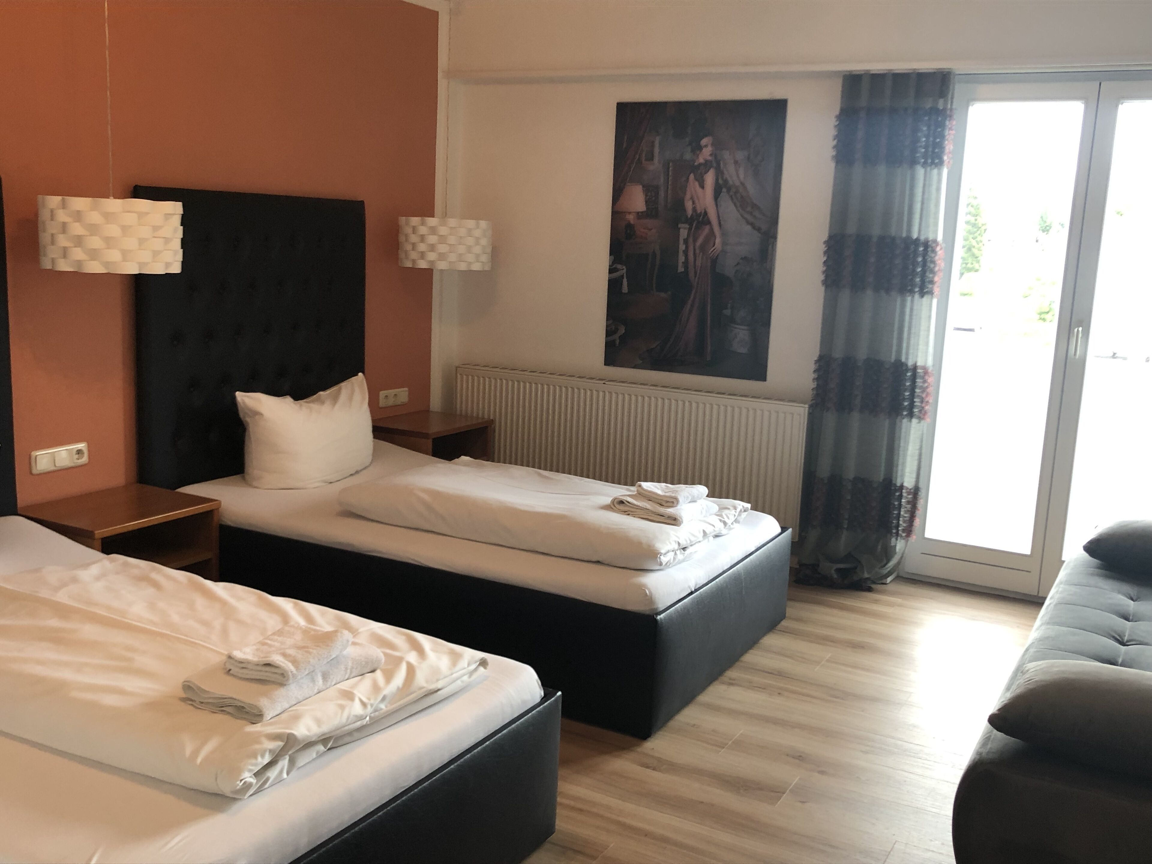 A hotel room with two beds, a large window with curtains, a radiator, and a painting on the wall.