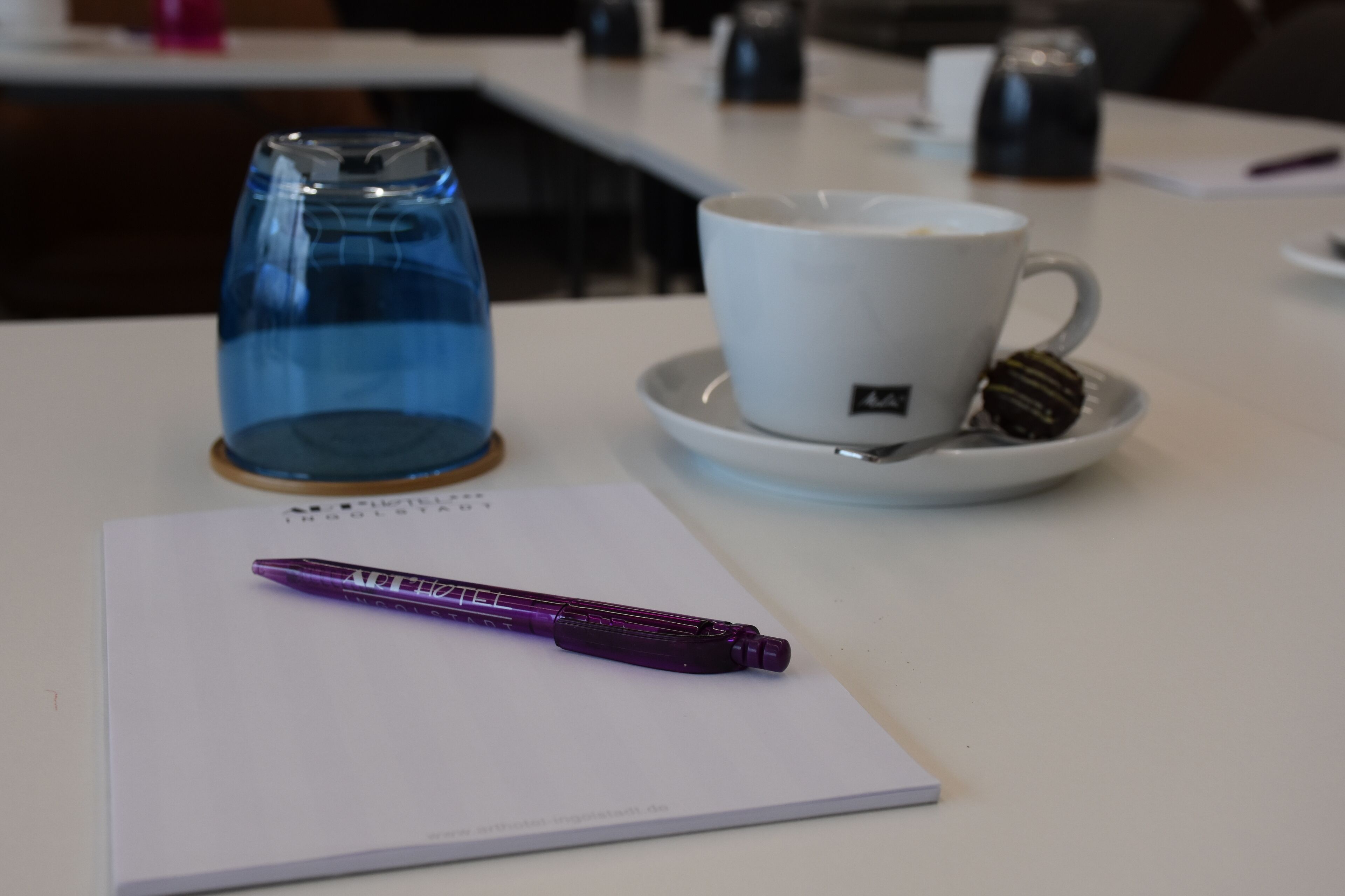 A white table with a purple pen, a white cup with a saucer, and a blue glass.