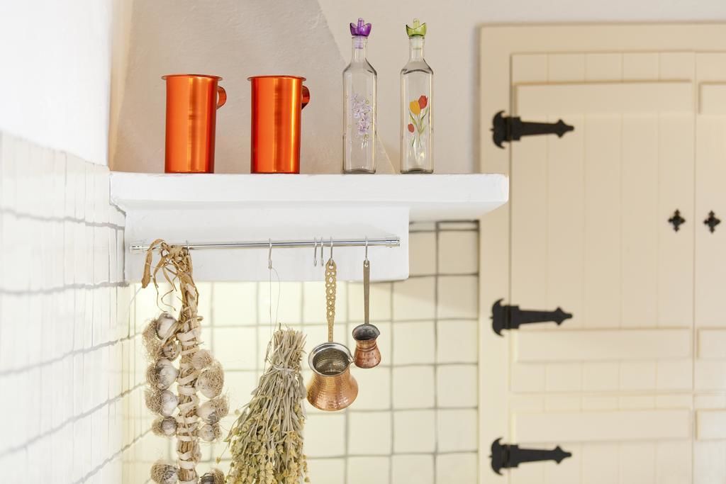 A kitchen with a white shelf holding orange mugs, glass bottles with flowers, and hanging dried herbs.