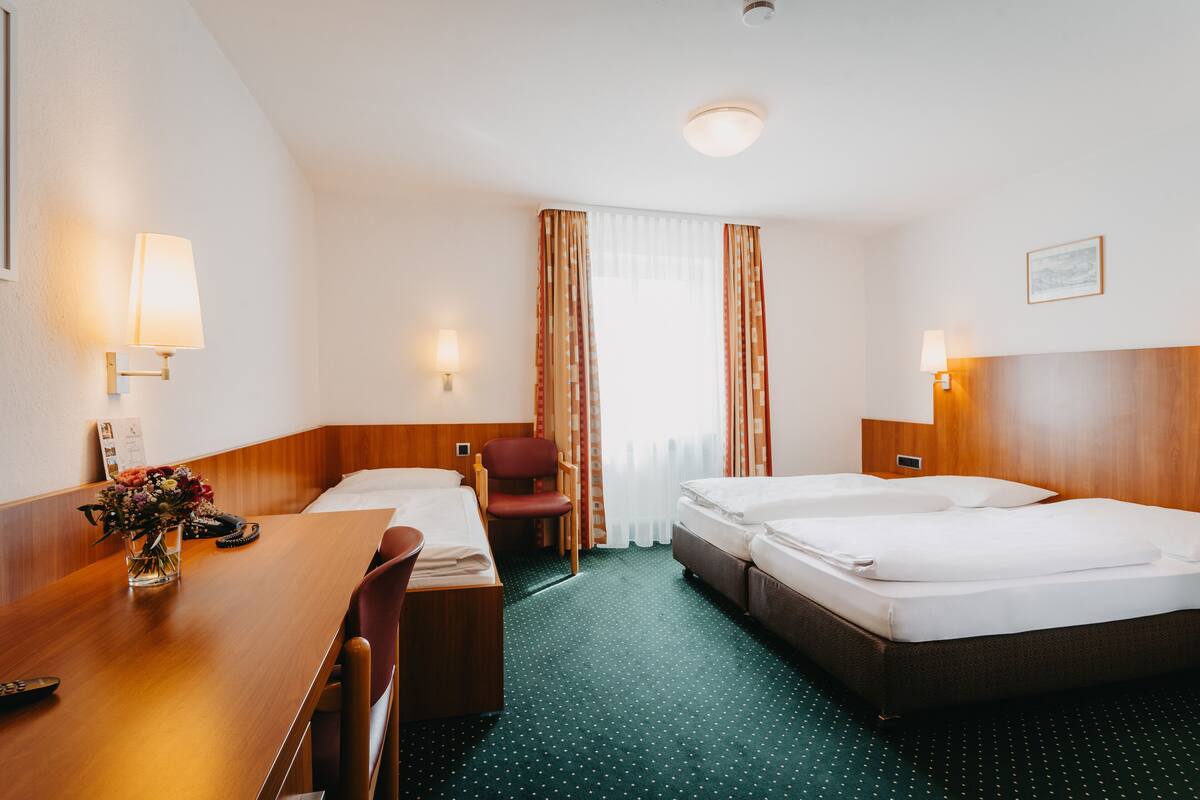 Habitación de hotel con dos camas individuales, un escritorio de madera, una silla, una mesita con un jarrón de flores y una ventana con cortinas.