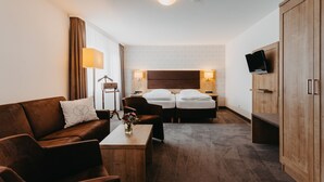 A hotel room with a brown leather sofa, a wooden coffee table with a vase, two single beds with white linens, a wooden wardrobe, and a flat-screen TV mounted on the wall. - Hirschenwirt (Eichstaett)