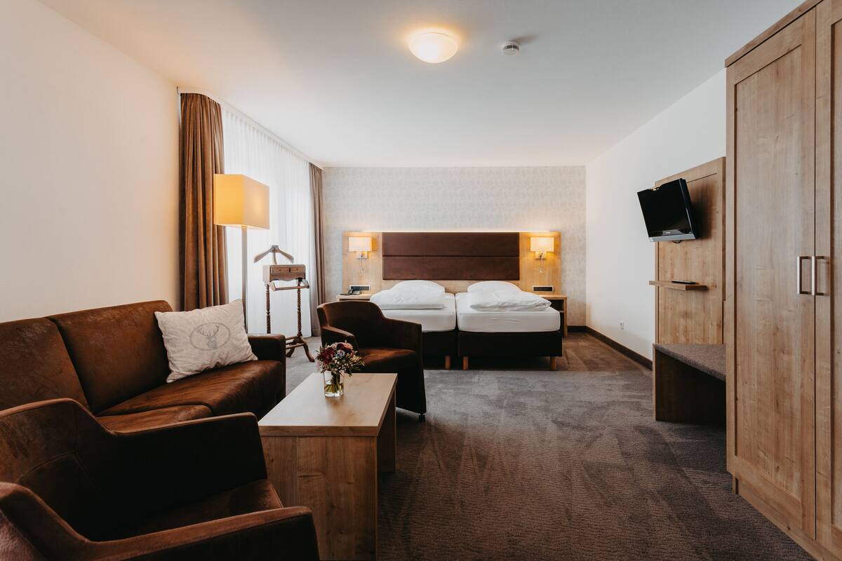Habitación de hotel con sofá de cuero marrón, una mesa de centro de madera con un florero, dos camas individuales con sábanas blancas, un armario de madera y un televisor de pantalla plana montado en la pared.