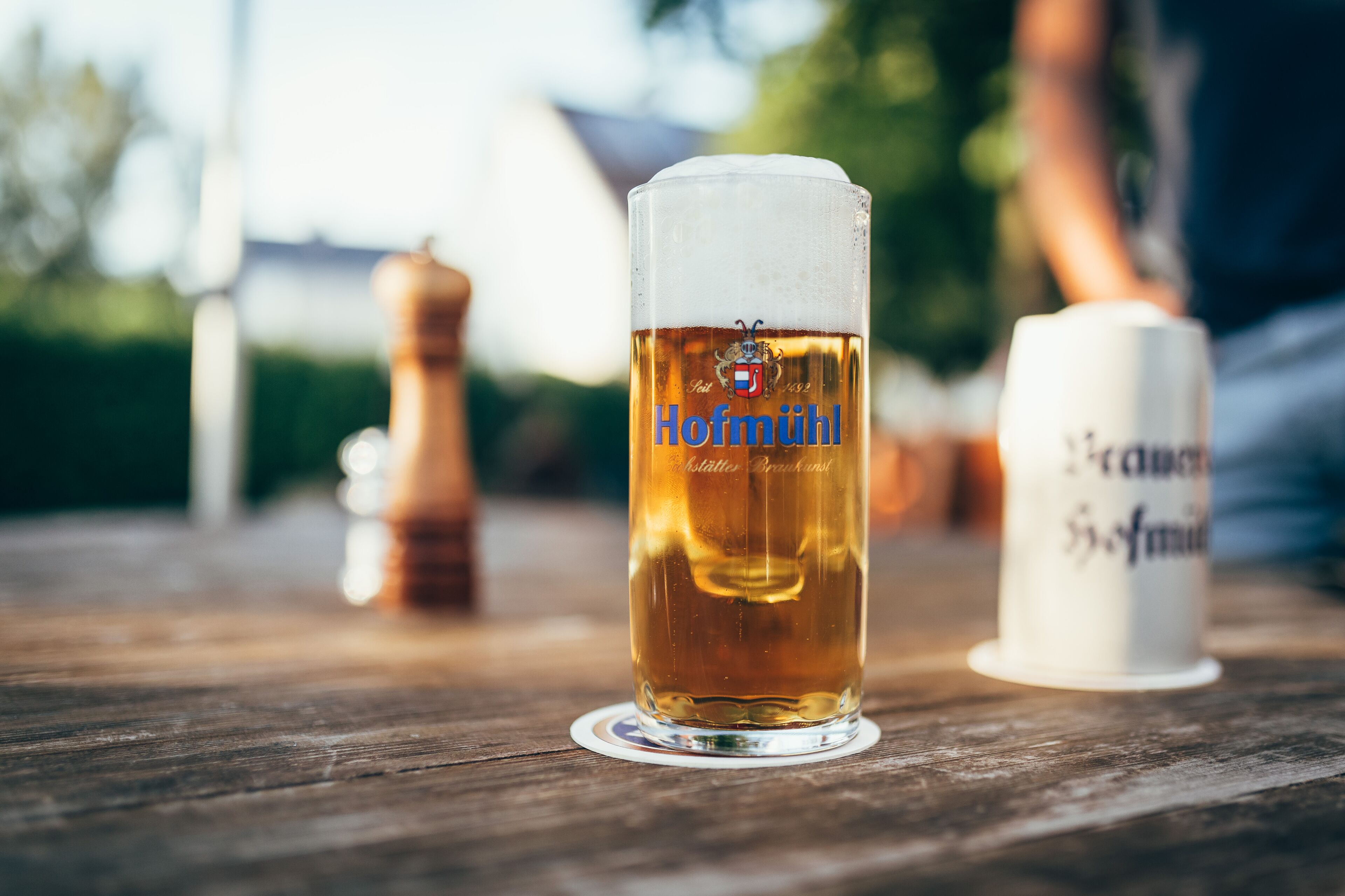 Um copo de cerveja com a inscrição "Hofmühl", apoiado em uma mesa de madeira com um moinho de pimenta e um saleiro por perto.