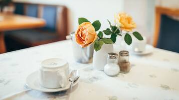 Una mesa con mantel floral, una taza y platillo blancos, una cuchara, un florero con dos rosas y saleros y pimenteros.