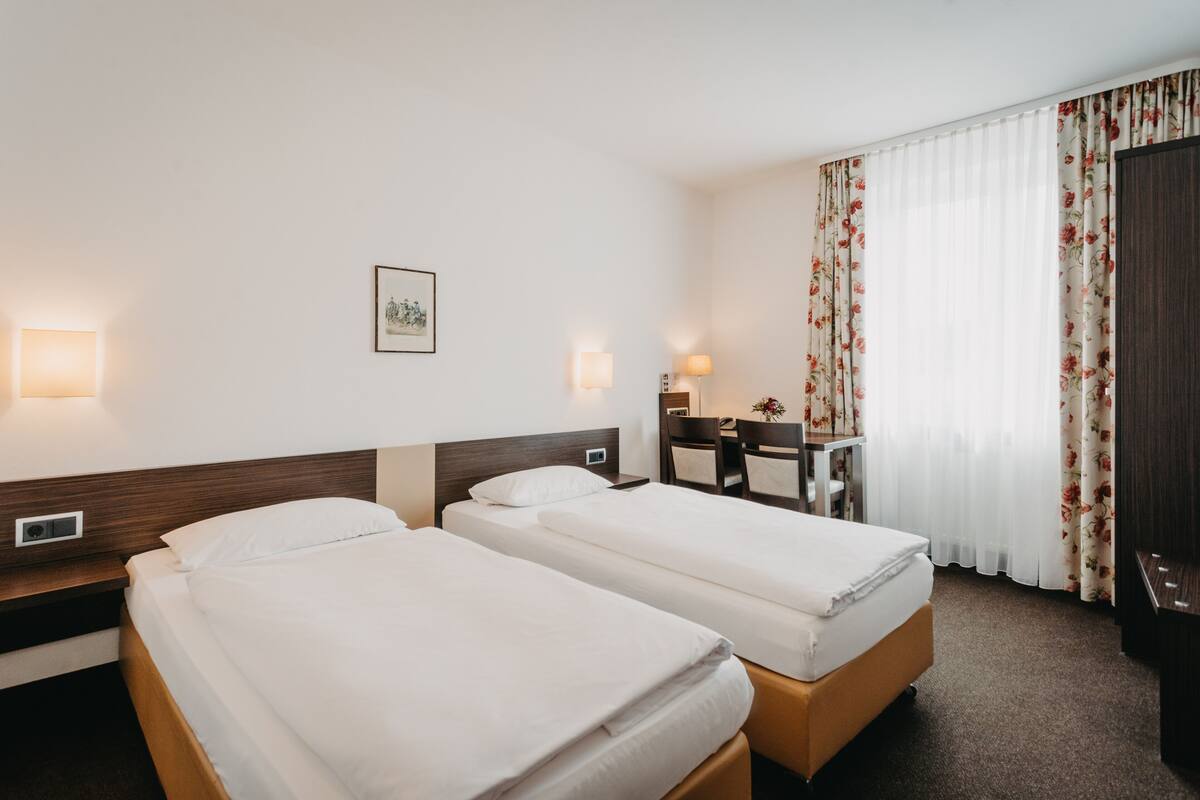 Habitación de hotel con dos camas individuales, un escritorio con silla, una mesita de noche con lámpara y un amplio ventanal con cortinas florales.