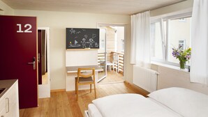 Et hotelværelse med en seng, et skrivebord, en stol, en rød dør med nummer 12 og et vindue med udsigt udad.