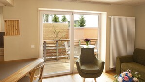 Et værelse med et træbord, en grøn lænestol, en sofa og udsigt gennem en skydedør til en terrasse.