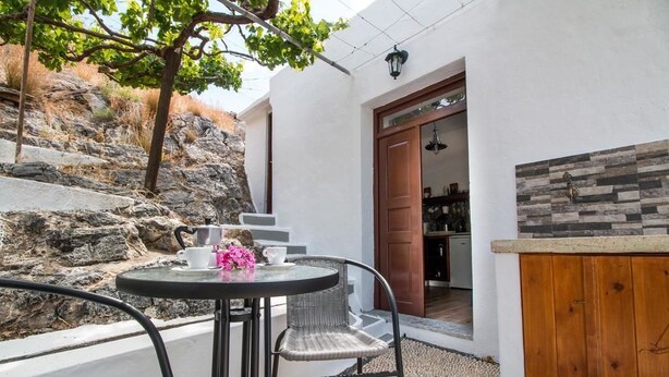 A patio with a round table and chairs, a wooden door leading to a kitchen, and a stone wall. - CasaLindos Spitaki (Rhodes)