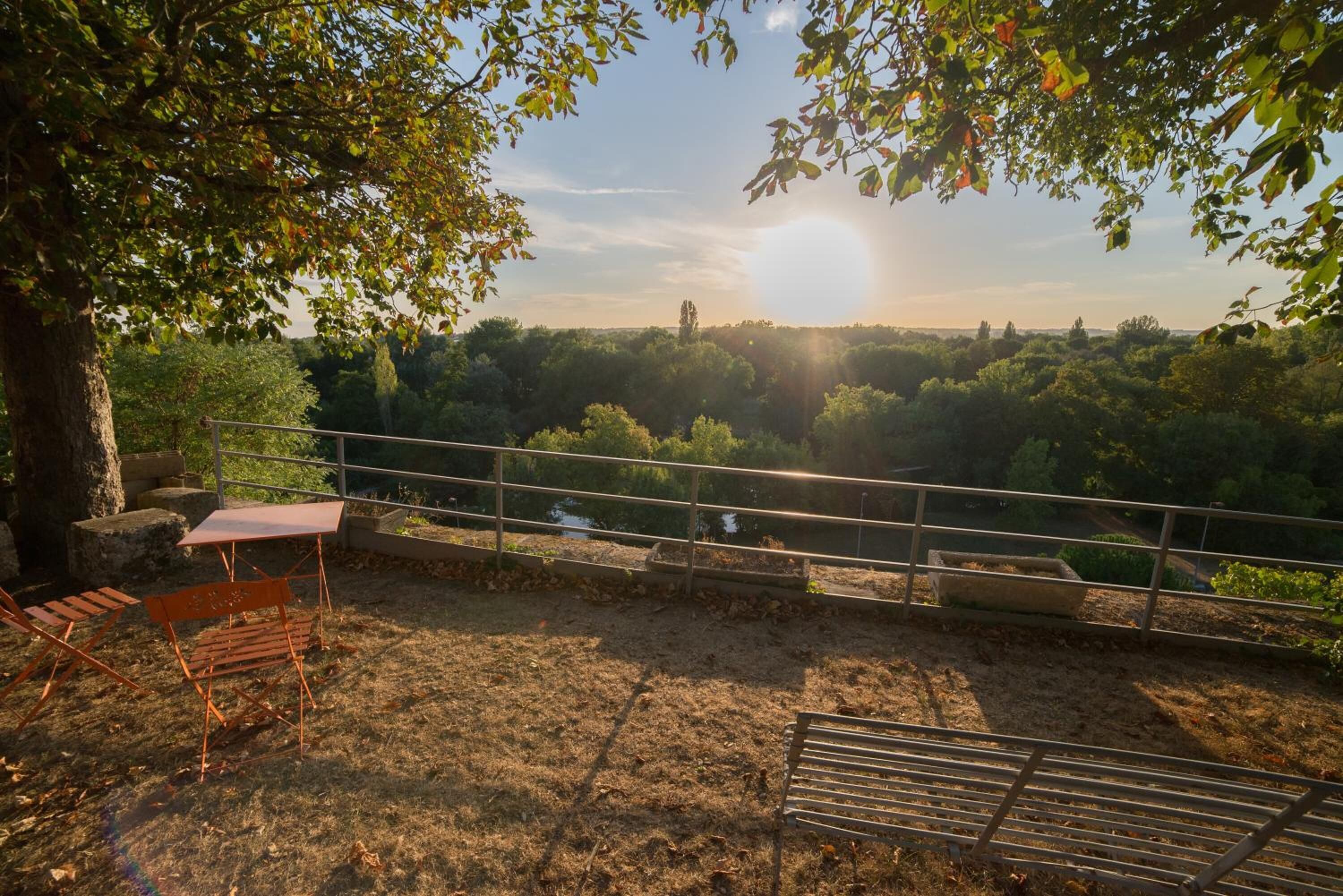 A terrace with a table and chairs, overlooking a forested area with a clear view of the sun setting behind the trees.