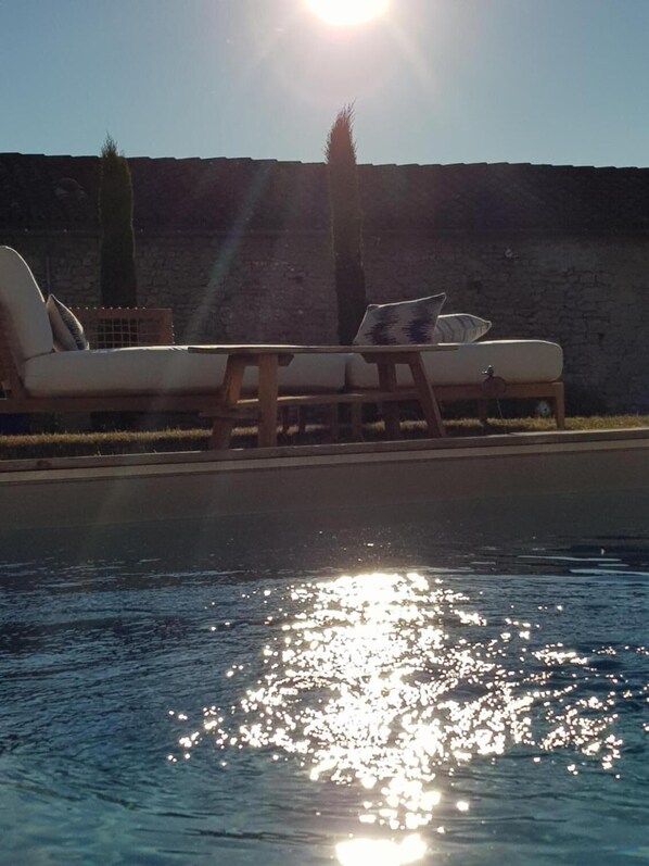 Un área junto a la piscina con dos sillas de descanso y una pequeña mesa, un muro de piedra al fondo, y el sol brillando sobre el agua.