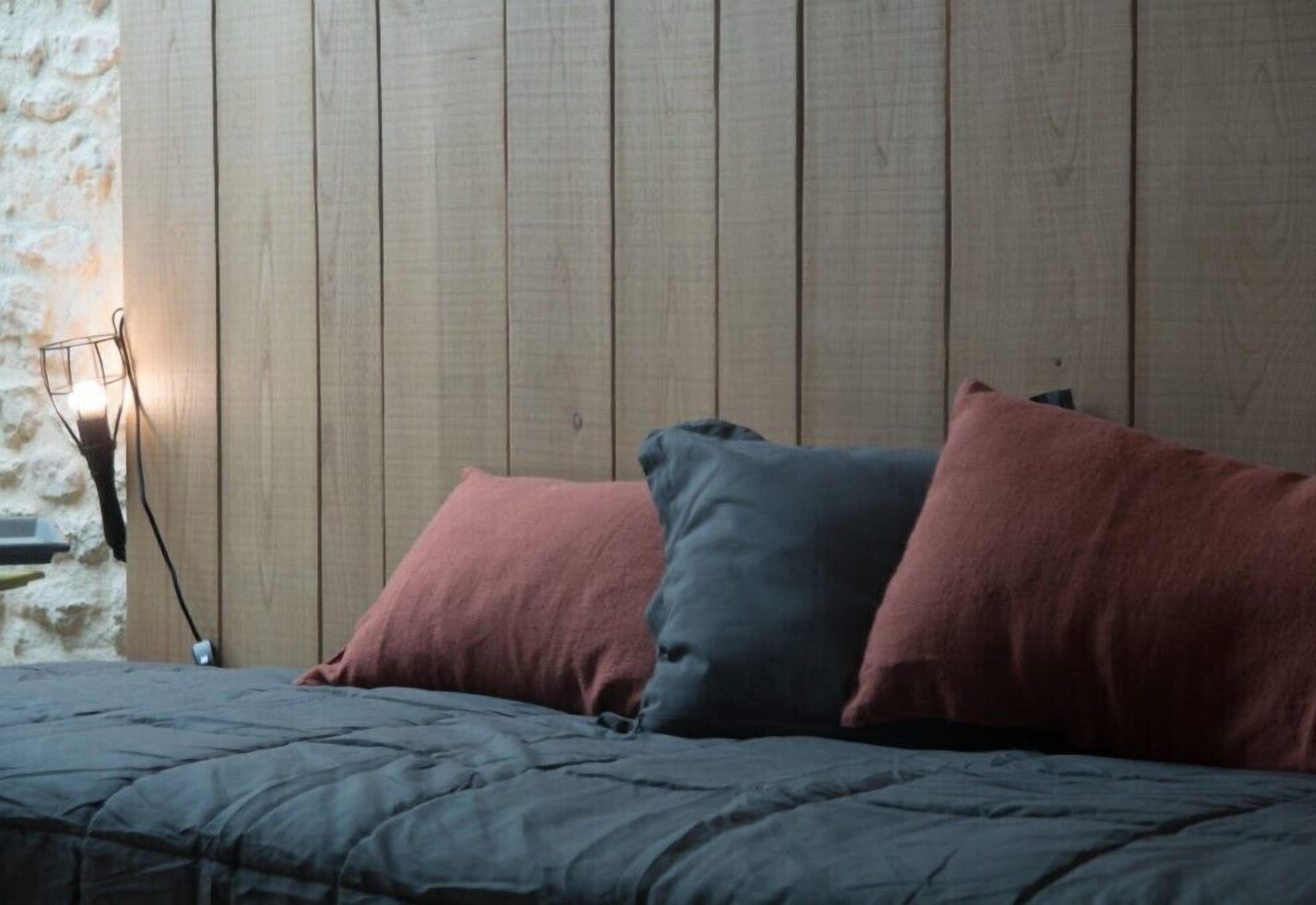 Een bed met een donkergrijze dekbedovertrek en twee kussens, een roze en een blauw, tegen een houten wand met een stenen element aan de linkerkant.