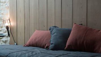 Een bed met een donkergrijze dekbedovertrek en twee kussens, een roze en een blauw, tegen een houten wand met een stenen element aan de linkerkant.