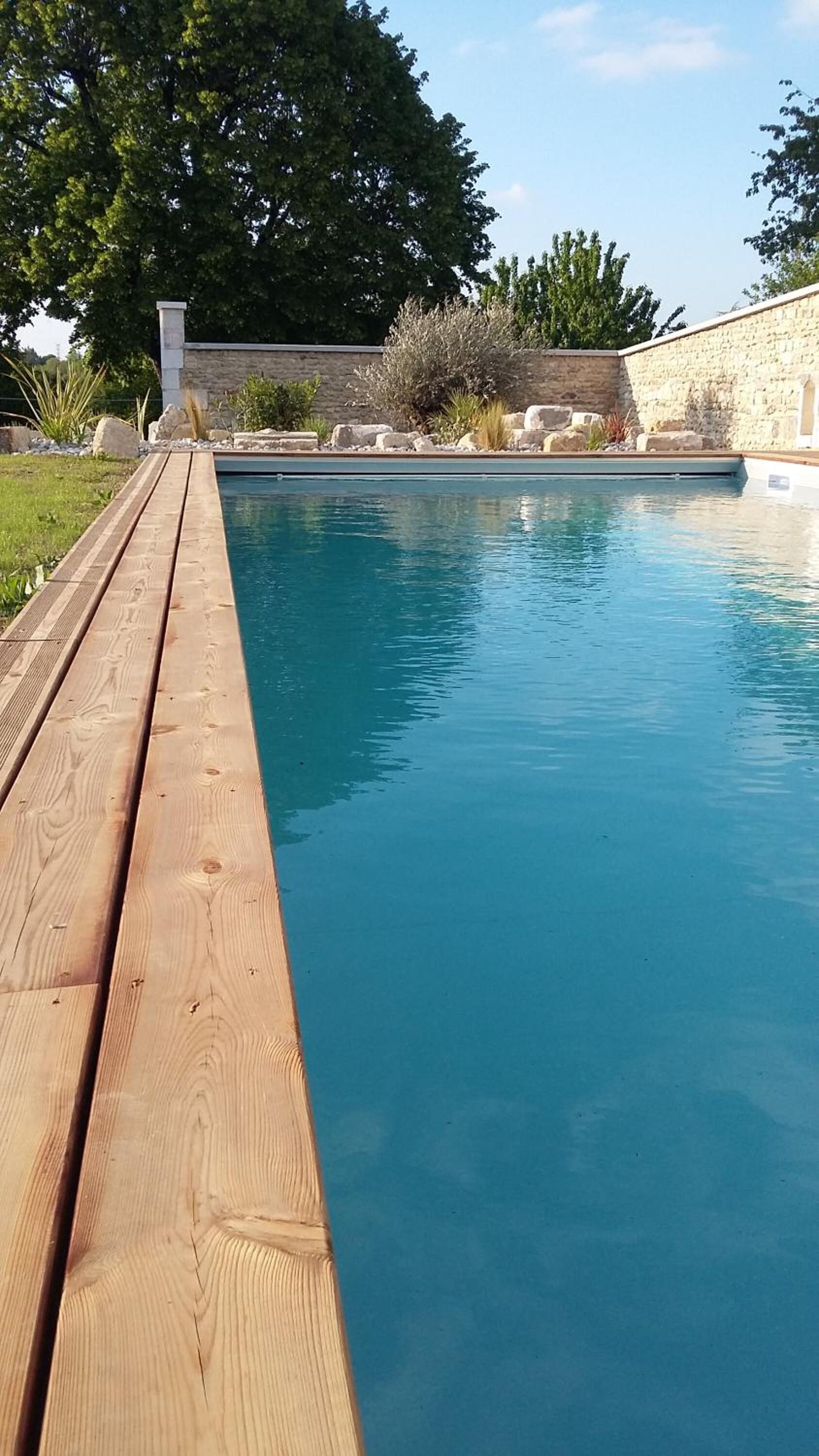 Um deck de madeira que leva a uma piscina retangular, com um muro de pedra e vegetação ao fundo.