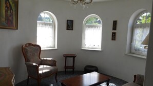 Una habitación con una araña, dos ventanas arqueadas, una silla de madera, una mesita y un sofá.